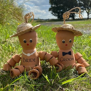 May include: Two terracotta pot figures dressed as a bride and groom. The bride wears a straw hat and a pearl necklace, and the groom wears a straw hat and a black bow tie. Both figures have painted faces and are sitting in a grassy area.