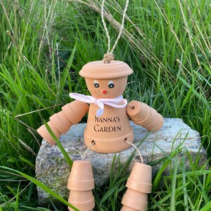 May include: A terracotta garden gnome decoration made from stacked terracotta pots. The gnome has a pink ribbon around its neck and a painted face. The gnome is sitting on a rock and has the words "Nanna's Garden" written on its torso.