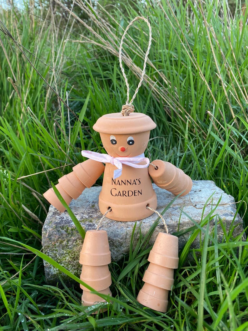 May include: A terracotta garden gnome decoration made from stacked terracotta pots. The gnome has a pink ribbon around its neck and a painted face. The gnome is sitting on a rock and has the words "Nanna's Garden" written on its torso.