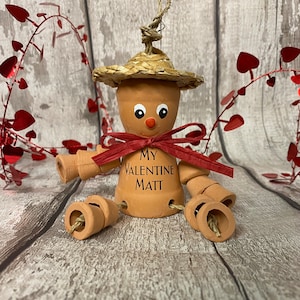 May include: A terracotta figurine with a straw hat, red bow, and the words "My Valentine Matt" written on the front. The figurine has a whimsical, handmade look and is perfect for a Valentine's Day gift.