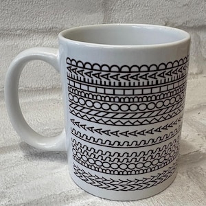 May include: White ceramic mug with a black geometric pattern. The design features various shapes and lines, including triangles, circles, and zigzags. The mug has a curved handle and is suitable for hot beverages.
