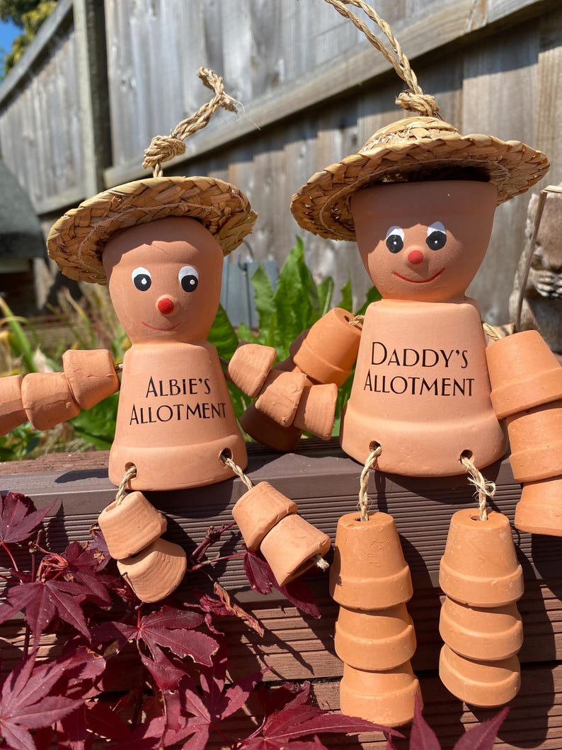 May include: Two terracotta pot figures with straw hats and rope hair. One reads "Albie's Allotment" and the other "Daddy's Allotment". The figures have painted eyes and red noses, with pot arms and legs.