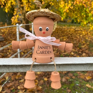 A terracotta garden pot person with a straw hat, pink ribbon, and the words 'Annie's Garden' written on its body. Can be customized with name of your choice.