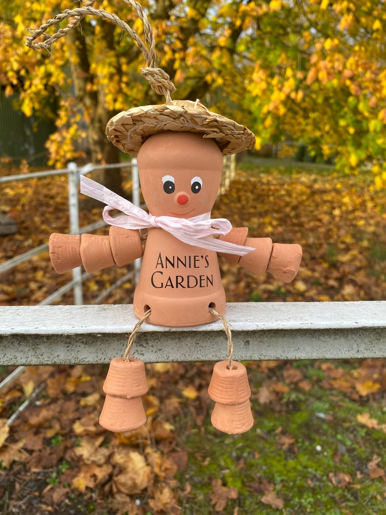 A terracotta garden pot person with a straw hat, pink ribbon, and the words 'Annie's Garden' written on its body. Can be customized with name of your choice.