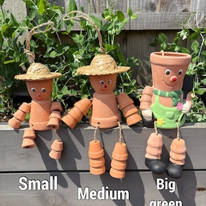 May include: Three terracotta pot people with straw hats and dangling pot feet. The figures are labeled "Small", "Medium", and "Big green". The "Big green" figure has a green apron and scarf. The background includes green foliage and a wooden fence.