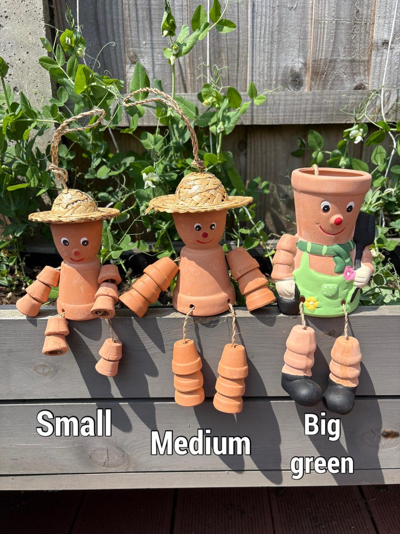 May include: Three terracotta pot people with straw hats and dangling pot feet. The figures are labeled "Small", "Medium", and "Big green". The "Big green" figure has a green apron and scarf. The background includes green foliage and a wooden fence.