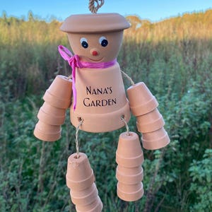 May include: A terracotta wind chime shaped like a person with a pink ribbon around its neck. The chime has a smiling face and the words "Nana's Garden" written on its torso. The chime is made of stacked terracotta pots.