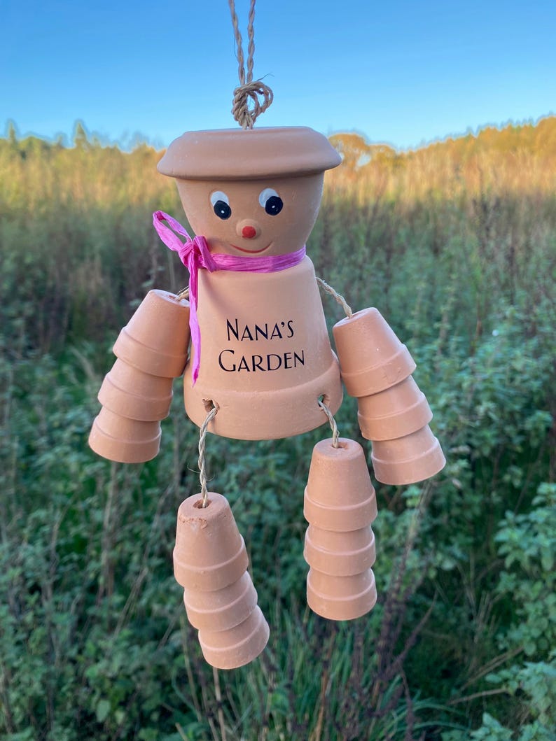 May include: A terracotta wind chime shaped like a person with a pink ribbon around its neck. The chime has a smiling face and the words "Nana's Garden" written on its torso. The chime is made of stacked terracotta pots.