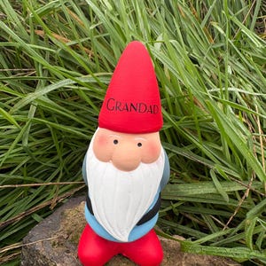 Op de afbeelding: Een rode en witte tuinkabouter met een rode muts waarop "Grandad" staat. De kabouter staat op een rots.