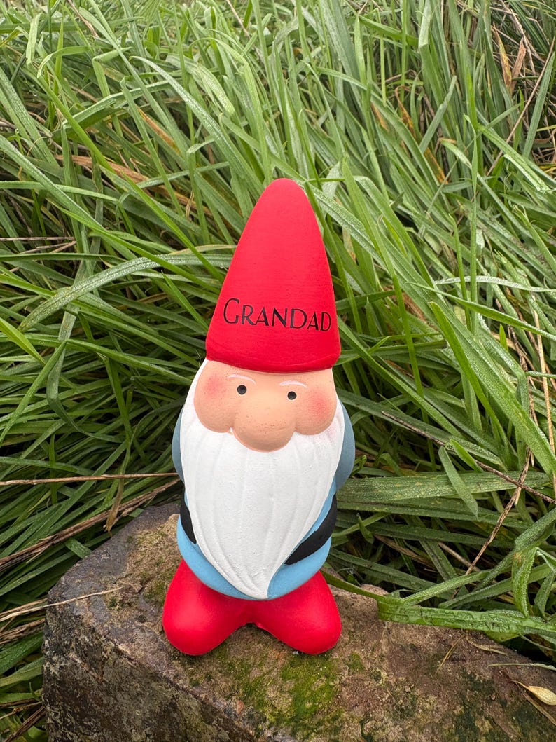 Op de afbeelding: Een rode en witte tuinkabouter met een rode muts waarop "Grandad" staat. De kabouter staat op een rots.