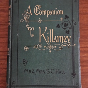 Puede incluir: Un libro verde vintage titulado "A Companion to Killarney" por Mr. & Mrs. S.C. Hall. El título y los nombres de los autores están en letras doradas. La portada tiene bordes negros decorativos y detalles florales.