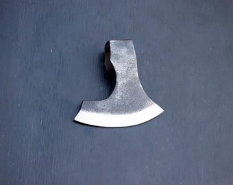 Peut inclure: Une petite hache forgée à la main avec une lame légèrement incurvée et une finition gris foncé. La tête de la hache est fixée à un manche en bois.