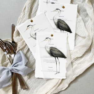 May include: Four white paper menus with a gray heron illustration and the word "MENU" in blue text. The menus are for an appetizer and dessert course. The menus are on a white linen tablecloth.