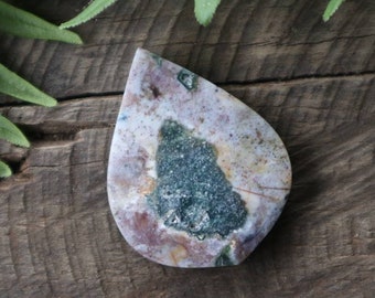May include: A teardrop-shaped, polished stone pendant with a white, pink, and brown base and green and black inclusions. The stone is set against a wooden background.