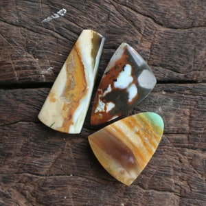 Op de afbeelding: Drie onregelmatig gevormde, gepolijste stenen met verschillende kleuren en patronen. De stenen zijn bruin, wit, groen en oranje. Ze zijn gerangschikt op een houten oppervlak.