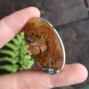 May include: A green, brown and white agate cabochon with a dendritic pattern. The cabochon is oval shaped and has a smooth, polished surface.