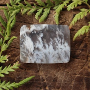 Puede incluir: Una losa de ágata cuadrada, blanca y gris con un patrón de remolino y nubes. La losa está sentada sobre un trozo de madera desgastada.