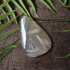 Könnte beinhalten: Ein weißer Achat-Cabochon mit durchscheinendem, wolkigen Aussehen und weißen und gelben Einschlüssen. Der Cabochon hat die Form einer Träne und eine glatte, polierte Oberfläche.