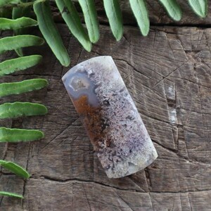 Könnte beinhalten: Ein polierter, rechteckiger Achat-Cabochon mit einem weißen, braunen und violetten Farbmuster. Der Achat hat eine glatte, polierte Oberfläche und eine natürliche, organische Form.