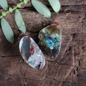 Könnte beinhalten: Zwei unregelmäßig geformte, polierte Steine mit einer Mischung aus roten, blauen, grünen und braunen Farben. Die Steine liegen auf einer dunkelbraunen Holzoberfläche.