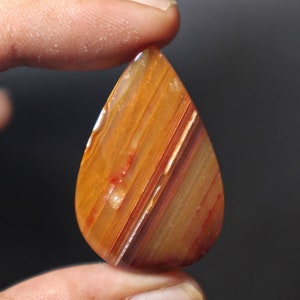 Peut inclure: Un cabochon d'agate en forme de larme avec un motif en couches d'orange, rouge et brun. La pierre est translucide et a une finition brillante.