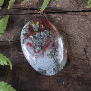 Peut inclure: Un pendentif ovale lisse en agate avec un motif blanc, gris et rouge. La pierre a un aspect naturel et organique avec une touche d'éclat.