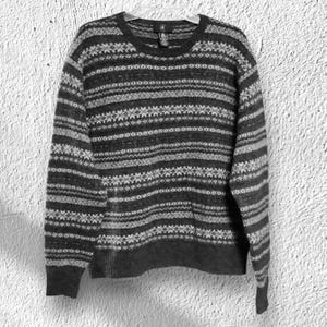 May include: A gray and white patterned sweater with a crew neck. The sweater features horizontal stripes and geometric designs. The long-sleeved sweater is hanging on a white hanger against a white textured background.