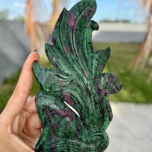 May include: A carved ruby in zoisite gemstone sculpture shaped like a stylized bird with flowing plumage. The stone features a deep forest green base color with prominent, irregular patches of magenta and deep red throughout the surface.