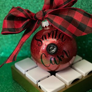 May include: A red glitter Christmas ornament with a black bow and the words "Santa Cam" written on it. The ornament is sitting on a gold stand with white squares.
