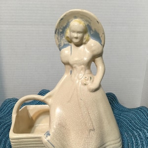 May include: Vintage ceramic figurine of a woman in a dress and hat, holding a basket. The figurine is cream-colored with blue accents on the hat and dress. The woman's dress has a floral detail. The figurine has a crackle finish.