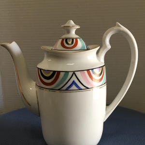 May include: White ceramic teapot with a colorful geometric design. The teapot has a lid with a gold trim and a handle.