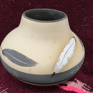 May include: A small, cream-colored ceramic vase with a gray rim and gray painted feathers. The vase has a white feather painted on the right side.
