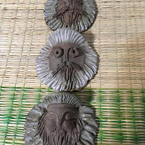 May include: Three ceramic face sculptures with detailed features. Each has a brown face, a long beard, and a textured, gray-white mane. The sculptures are arranged vertically against a woven background.
