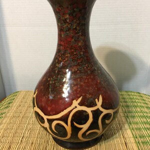 May include: A decorative ceramic vase with a flared top and a bulbous body. The vase features a speckled red and brown glaze with a cream-colored, swirling design near the base. The vase sits on a dark brown base.
