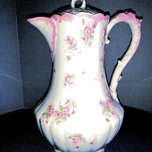 May include: A white porcelain teapot with a pink floral pattern and a pink and silver lid. The teapot has a fluted body and a curved handle.