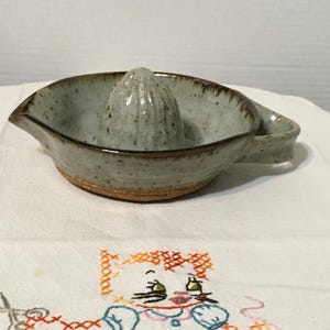 May include: A ceramic citrus juicer with a light blue-grey glaze and brown trim. The juicer has a built-in reamer and a pouring spout. The handle is a small loop. The juicer is on a white surface.