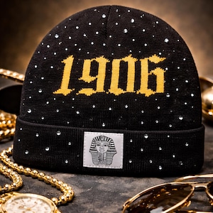 May include: Black knit beanie with the number "1906" in gold lettering and scattered rhinestones. A small rectangular patch features a pharaoh graphic. Gold jewelry and sunglasses are in the background.