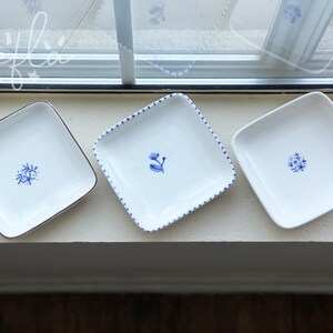 May include: Three white square ceramic dishes with blue floral designs. The dishes are arranged in a row on a white surface.