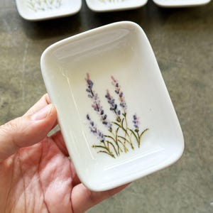 May include: A set of small, white, rectangular ceramic dishes. Each dish features a hand-painted lavender sprig design in purple and green. The dishes are held in a hand, with the word "leefla" in cursive script at the bottom.