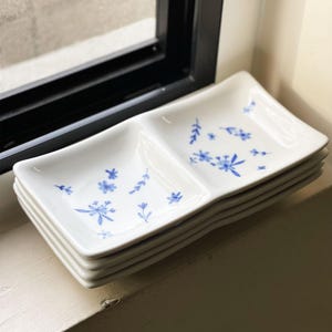 May include: A stack of four white ceramic divided serving dishes with a blue floral pattern. The dishes are stacked on a white surface.
