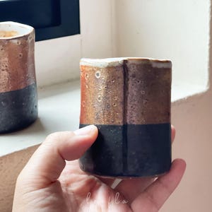 May include: Two handmade ceramic tumblers. The top half of each tumbler is a mottled brown, while the bottom half is a solid dark blue. The tumblers have a rustic, textured appearance, suggesting a handcrafted origin.