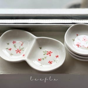 May include: White ceramic serving dishes with a two-section design, adorned with delicate pink floral patterns and green leaves. The dishes are stacked, showcasing the design. The word "leefla" is written in cursive.