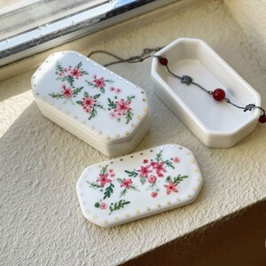 May include: Three white ceramic trinket boxes with floral designs and gold accents. One box is open, revealing a silver necklace with red beads. The boxes are arranged on a light-colored surface, with sunlight casting shadows.