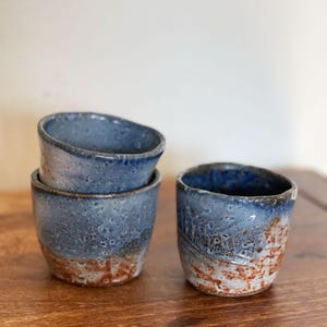 May include: Three small ceramic cups with a blue and white glaze. The cups have a rustic, handmade look and are stacked on top of each other.
