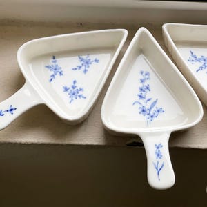 May include: Three white ceramic triangle-shaped dishes with blue floral designs. The dishes have handles and are perfect for serving small snacks or appetizers.