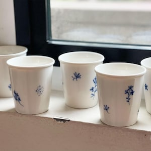 May include: A set of five white ceramic cups, each featuring small, hand-painted blue floral and botanical motifs scattered across the exterior surface. The cups have a simple, cylindrical shape with a slightly flared rim and a smooth finish.