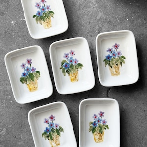 May include: Six white rectangular ceramic dishes, each adorned with a hand-painted floral design. The design features blue and purple flowers in a yellow pot. The dishes are arranged on a dark gray surface.