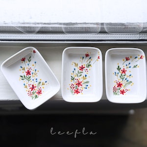 May include: Three white rectangular dishes with rounded corners, each adorned with a floral design. The flowers are red, with yellow and blue accents, and green leaves. The dishes are arranged on a white surface.
