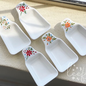 May include: Five white ceramic serving dishes with floral designs. Each dish has a handle and is shaped like a small boat. The designs feature red, orange, blue, and green flowers.