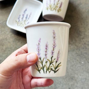 May include: White ceramic cup with a hand-painted lavender design. The cup features purple lavender flowers with green stems and leaves. Other matching ceramic pieces are visible in the background.